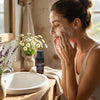 Woman applying foaming facial wash with chamomile and lavender near sink with flowers